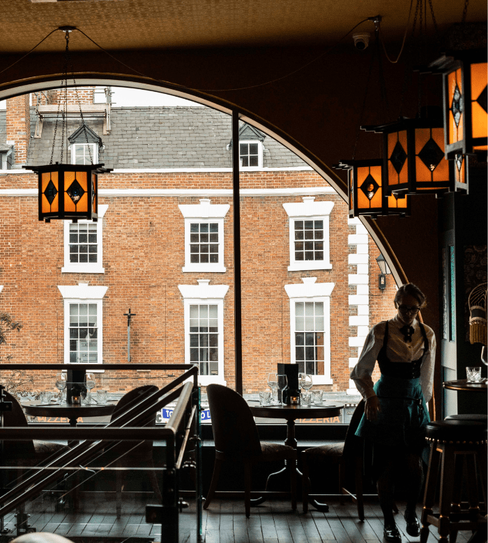 A server walks past tables inside a dimly lit café with large arched windows, through which brick buildings with white-trimmed windows are visible across the street. Orange pendant lamps hang from the ceiling.