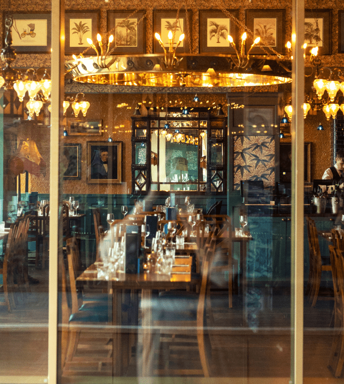 A view through a window into a warmly lit restaurant, showing set tables with glasses and napkins, chandeliers, framed art on the walls, and a bartender standing behind a counter.