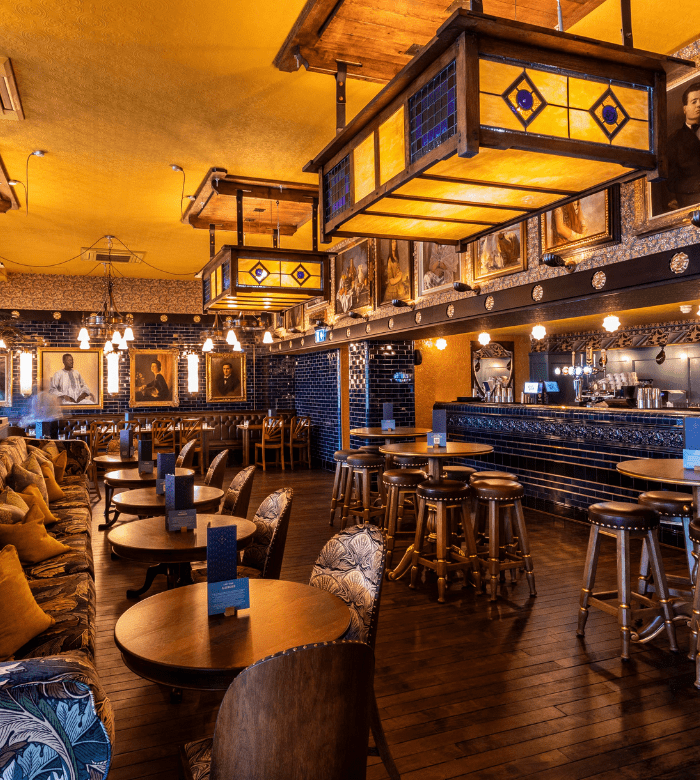 A warmly lit bar with wooden tables and stools, cushioned seating, large decorative lamps, framed portraits on the walls, and a polished wooden floor. The atmosphere is cozy and inviting.