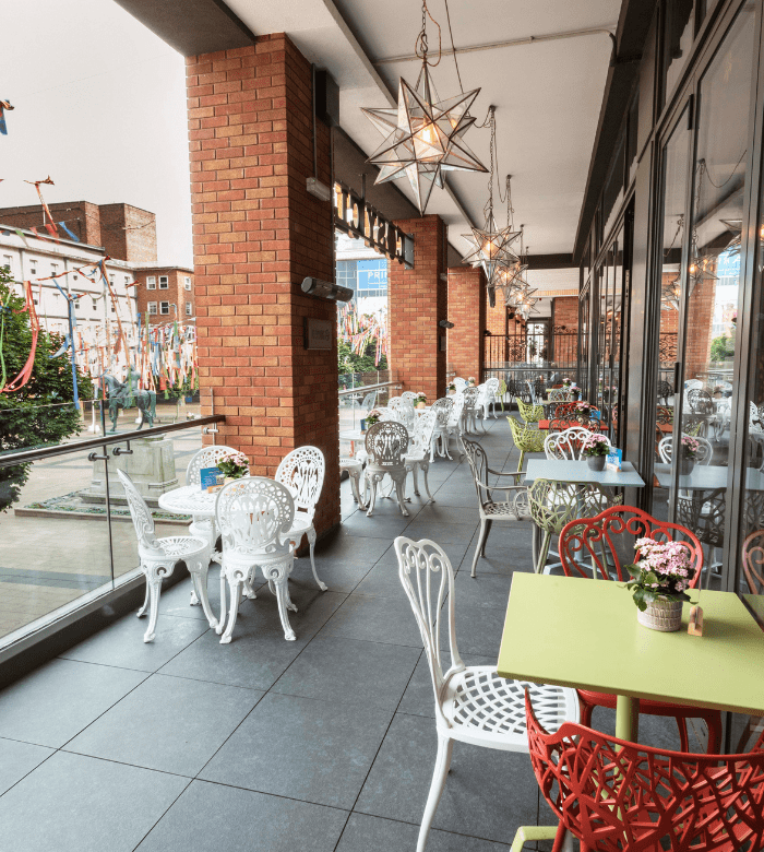 Outdoor cafe with colorful metal chairs and tables, potted flowers, and star-shaped hanging lights. The seating area is next to large brick columns and glass panels, overlooking a lively city street.