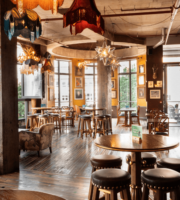 Cozy, sunlit café with wooden floors, round tables, cushioned stools, eclectic pendant lights, framed art on ochre walls, large windows, and leafy greenery visible outside.