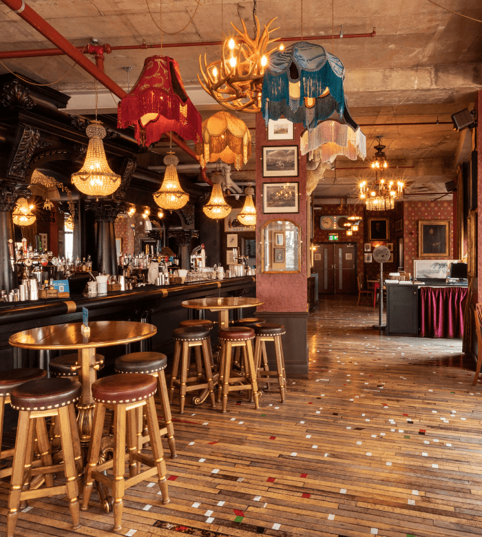 A vintage-style bar with ornate woodwork, colorful fringed lampshades, chandeliers, a polished wooden bar, tall stools, framed pictures on the wall, and a patterned wooden floor.