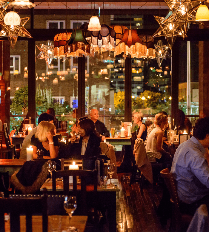 A warmly lit restaurant filled with people dining, talking, and enjoying drinks at candlelit tables. Decorative lamps and star-shaped lights hang from the ceiling, with city lights visible through large windows.
