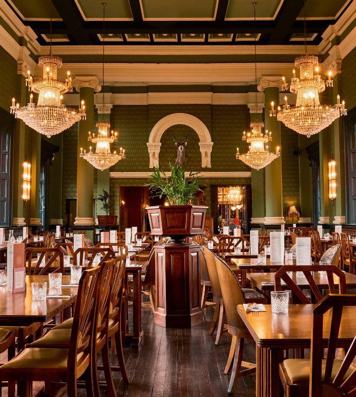 Elegant restaurant interior with wooden tables and chairs, ornate chandeliers, green walls, and a central plant arrangement. Large windows and an arched alcove with a statue enhance the luxurious, classic atmosphere.