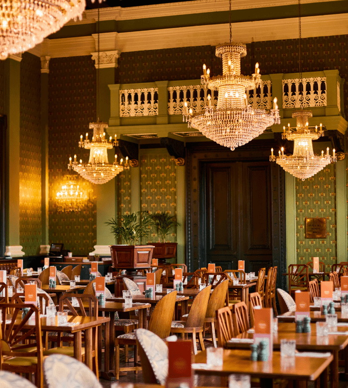 Elegant restaurant dining room with wooden tables and chairs, ornate chandeliers, decorative green walls, and tall double doors. The tables are set with glasses and menus, and warm light fills the space.