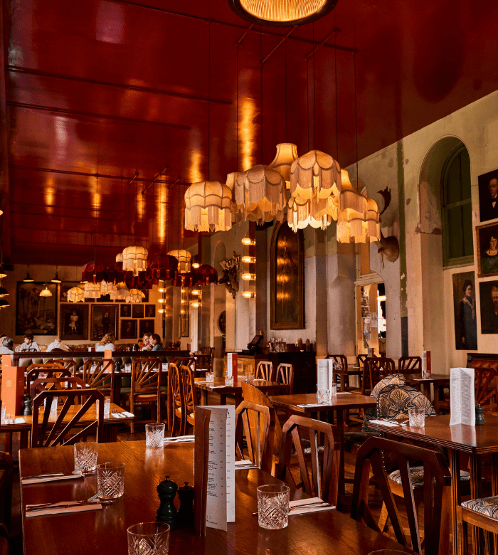 A warmly lit restaurant with wooden tables and chairs, vintage hanging lamps, glassware, and menus on tables. The ceiling is a deep red, and portraits and decorations adorn the walls, creating a cozy, eclectic atmosphere.