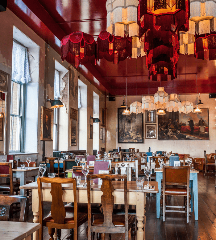A colorful restaurant interior with eclectic wooden tables and chairs, ornate lampshades hanging from a red ceiling, large windows with curtains, and framed paintings on the walls. Tables are set with glasses and cutlery.