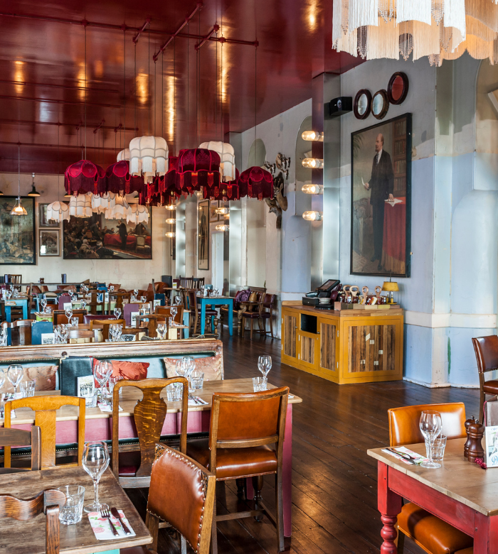 A vibrant restaurant with eclectic decor, featuring mismatched wooden chairs, set tables with glassware, colorful pendant lamps, wall art, and a large portrait on a pale blue wall with a shiny red ceiling.