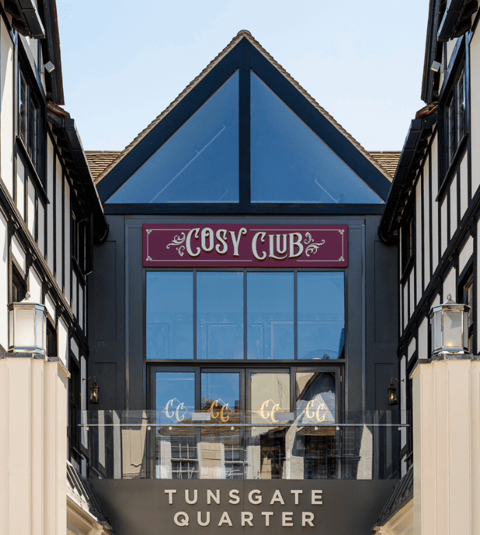 A modern building with black and white timber framing features a glass-fronted entrance. Signs read "Cosy Club" above and "Tunsgate Quarter" below, with reflections visible in the windows.