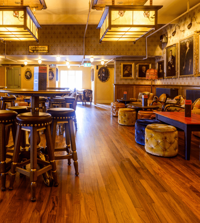 A cozy pub interior with wooden floors, high stools, plush ottomans, patterned wallpaper, and framed portraits on the wall. Warm lighting and sunlight from a window create a welcoming atmosphere.