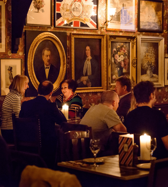 A group of people sit and converse around candlelit tables in a cozy, warmly lit restaurant with framed paintings covering the walls.