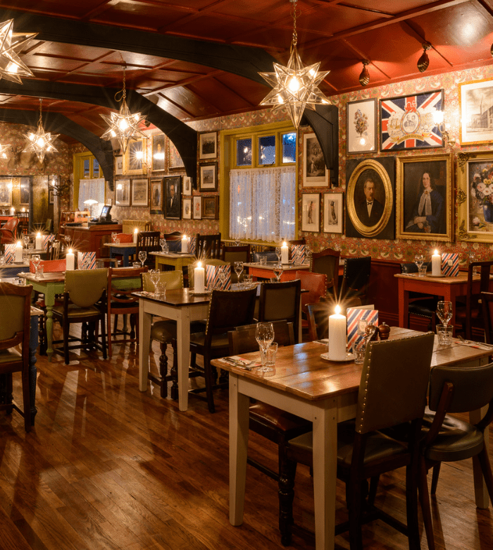 A cozy, warmly lit restaurant with wooden floors, candlelit tables, eclectic vintage chairs, and walls decorated with framed portraits, paintings, and British-themed decor. Star-shaped pendant lights hang from the ceiling.