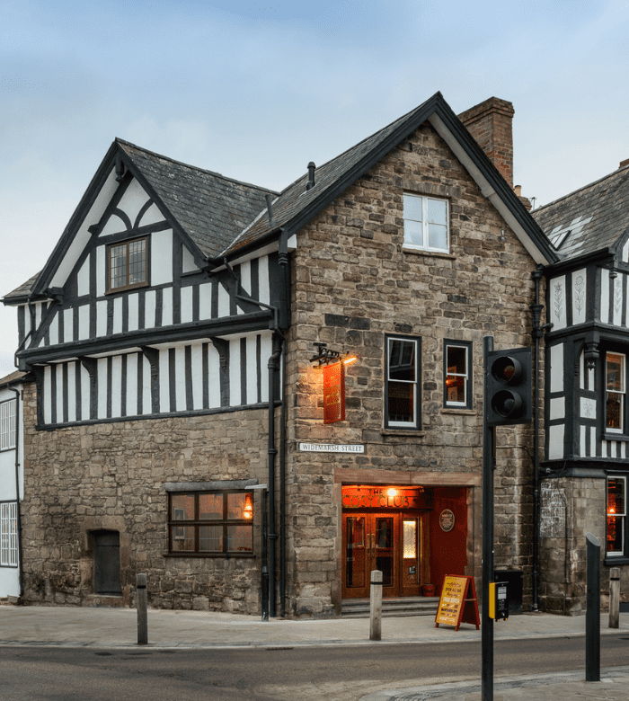 A historic stone and timber-framed pub with a red sign and warm lights glowing from its windows, located on a street corner with a traffic light and street sign nearby.