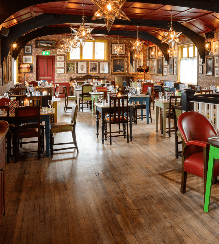 A cozy restaurant with wooden floors, colorful mismatched chairs, tables set for dining, star-shaped pendant lights, and walls decorated with framed pictures and artwork. Natural light streams through the windows.