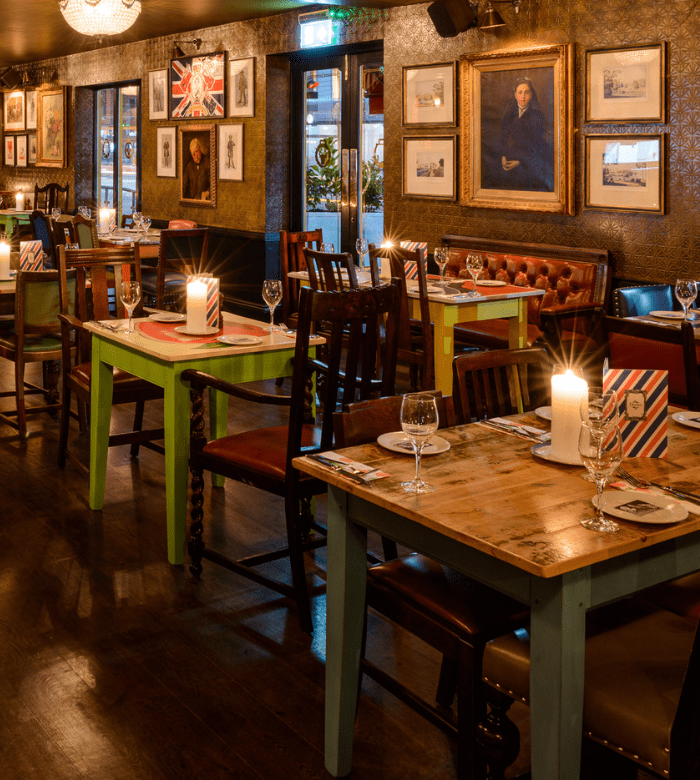 A cozy, warmly lit restaurant with mismatched wooden tables and chairs, candles on each table, framed art on the walls, and a relaxed, inviting atmosphere.