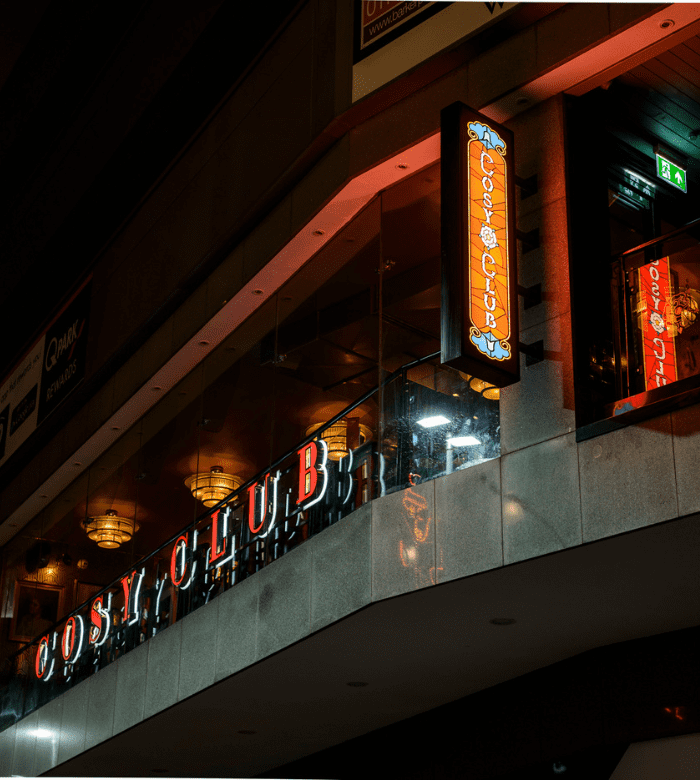 Nighttime photo of a building with an illuminated red and orange "COSY CLUB" sign on a balcony, featuring warm interior lighting and reflections of the sign in the glass panels.