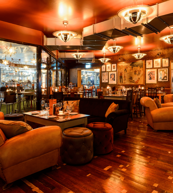 A cozy restaurant with warm lighting, plush armchairs, wooden floors, and art-covered walls. Tables are set for dining, and the space feels inviting and stylish with a relaxed, intimate atmosphere.