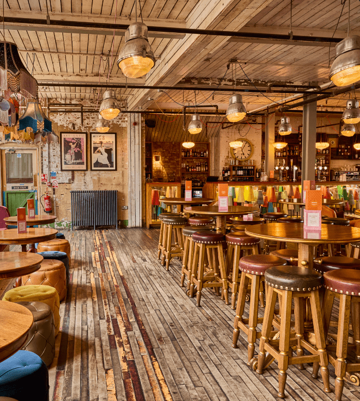 A spacious, rustic bar with wooden floors and ceiling, round wooden tables with stools, colorful decor, warm lighting, and a well-stocked bar in the background. Framed art and retro details add character to the cozy space.