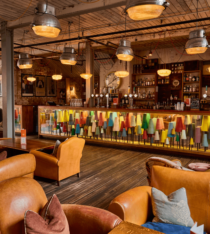A cozy bar with tan leather chairs, colorful lampshades lining the bar front, wooden floors, warm lighting, and shelves of bottles behind the bar, creating a welcoming, eclectic atmosphere.