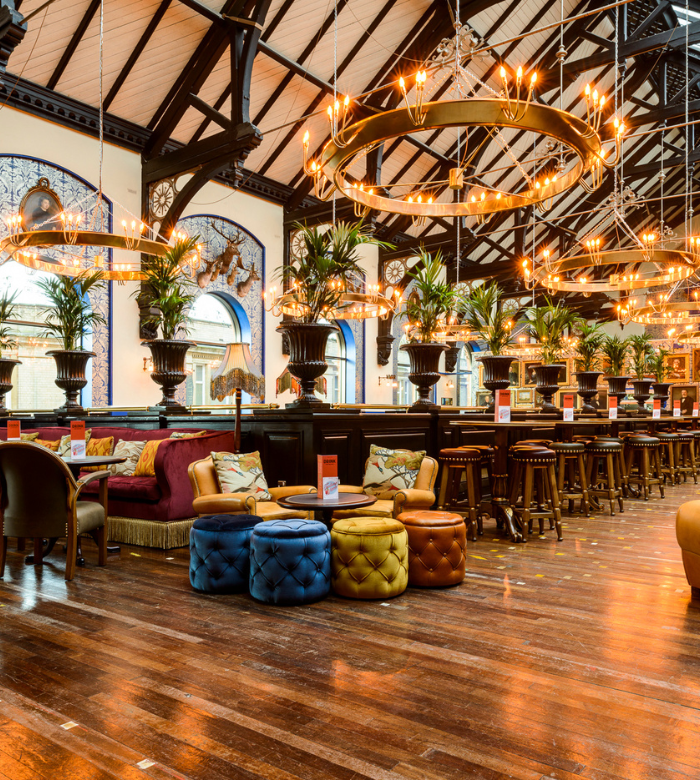 A spacious, stylish restaurant with wooden floors, tufted stools, plush seating, chandeliers, and tall plants. Large arched windows let in natural light, and the decor mixes vintage and modern elements.