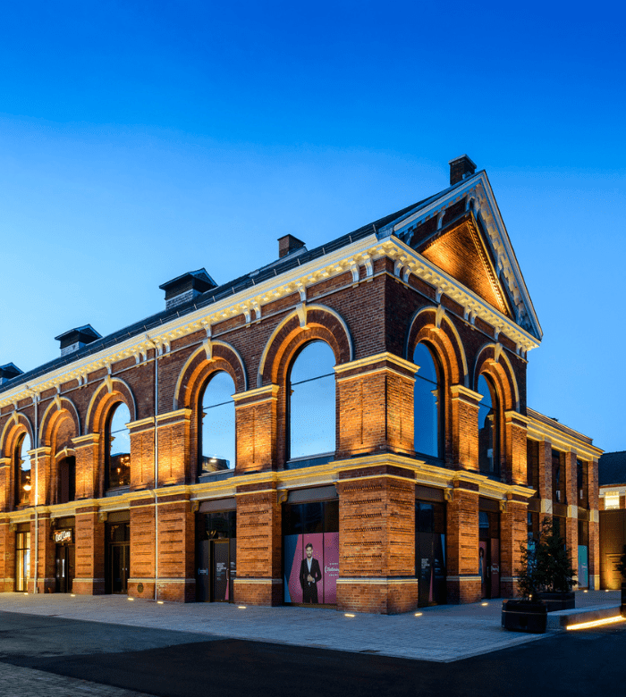 A historic brick building with large arched windows is illuminated by warm lights at dusk, set against a clear blue sky. The structure combines classic architectural details with modern lighting.