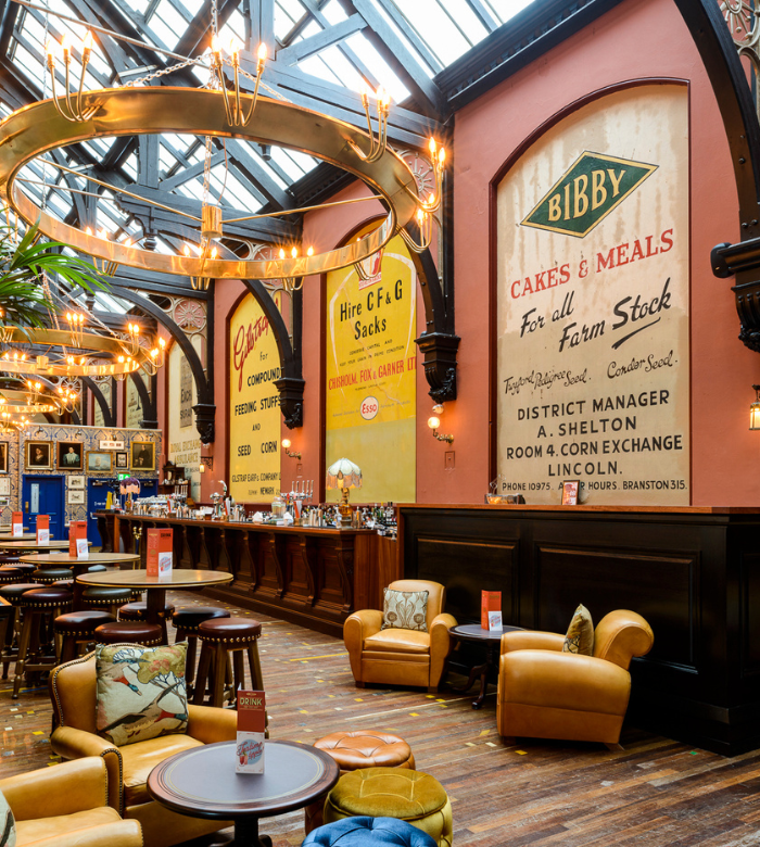 A spacious, stylish pub interior with plush chairs, wooden tables, a high glass ceiling, and large vintage wall ads. Warm lighting and a well-stocked bar create an inviting atmosphere.