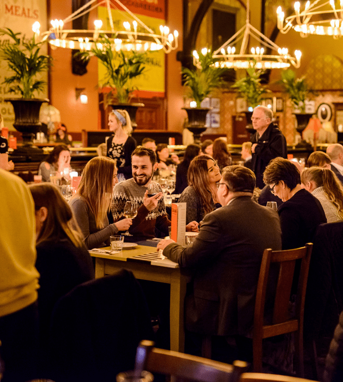 A lively restaurant scene with people sitting at tables, talking and laughing under warm chandeliers. Green plants decorate the space, and the atmosphere appears cozy and social.