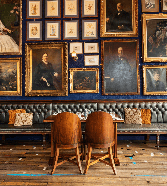 A cozy seating area with two wooden chairs and a tufted leather banquette is set against a wall covered in vintage portraits and framed artwork in gold and wood frames. The wooden floor has colorful tape markings.