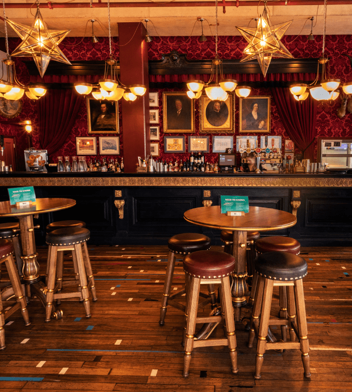 A vintage-style bar with ornate gold and red decor, round wooden tables and stools, hanging star-shaped lights, and framed portraits on the wall behind the bar counter.