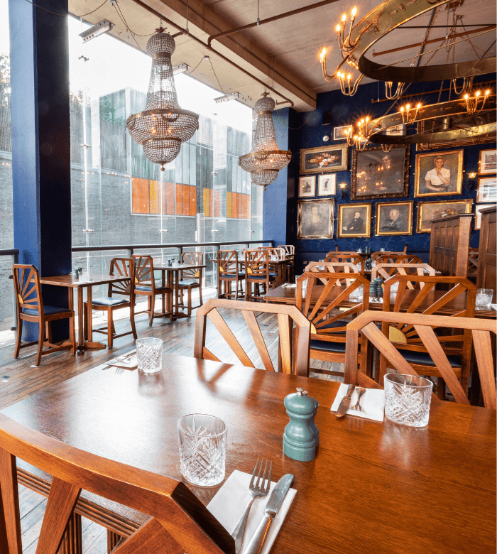 A stylish restaurant interior with wooden tables and chairs, crystal glasses, chandeliers, framed artwork on the wall, and large windows letting in natural light.