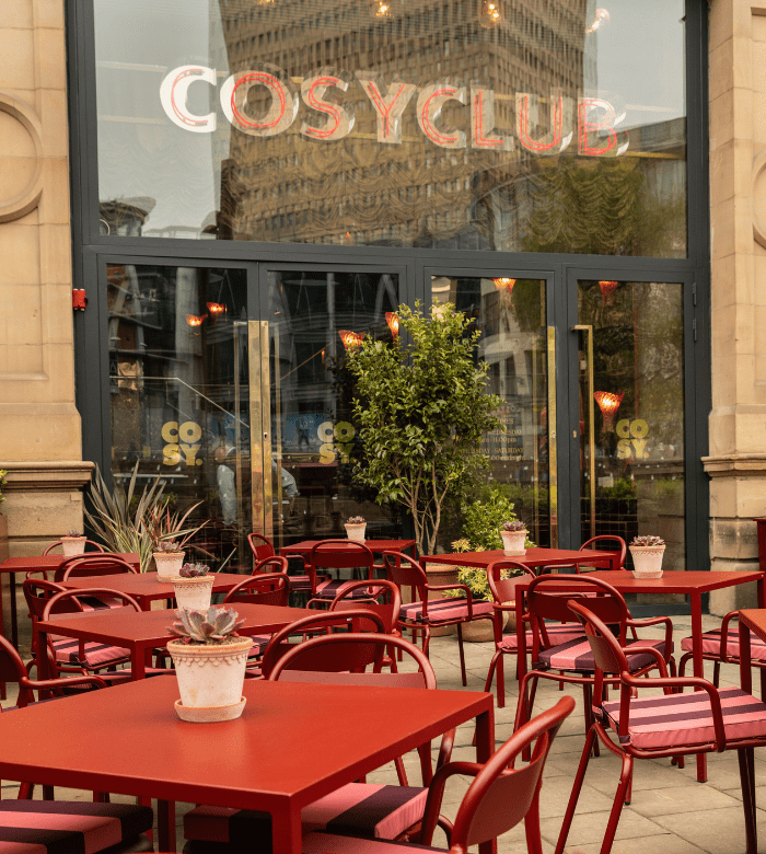 Outdoor seating area with red tables and chairs in front of a restaurant called "Cosy Club." Potted plants decorate the tables, and large glass doors reflect an urban cityscape in the background.