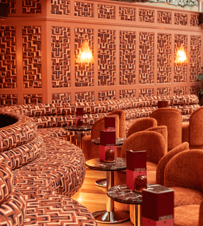 A stylish lounge with patterned, curved seating and round marble tables, each holding red menus and a small candle. The walls have geometric designs in shades of red and orange, and warm sconces provide soft lighting.