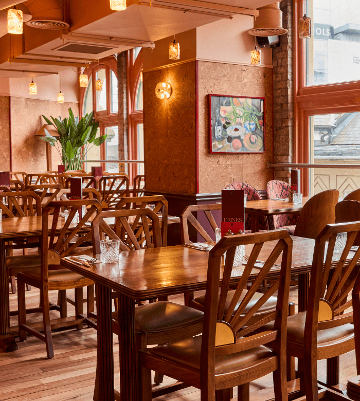 A cozy restaurant dining area with wooden tables and chairs, warm lighting, a plant in the corner, artwork on the wall, and large windows letting in natural light.