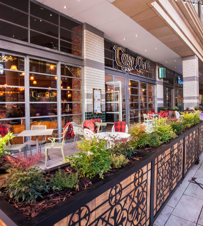 Outdoor seating area of a café with tables, chairs, and red blankets, separated from the sidewalk by a decorative planter filled with green plants; large windows and the café sign "Cosy Club" are visible.