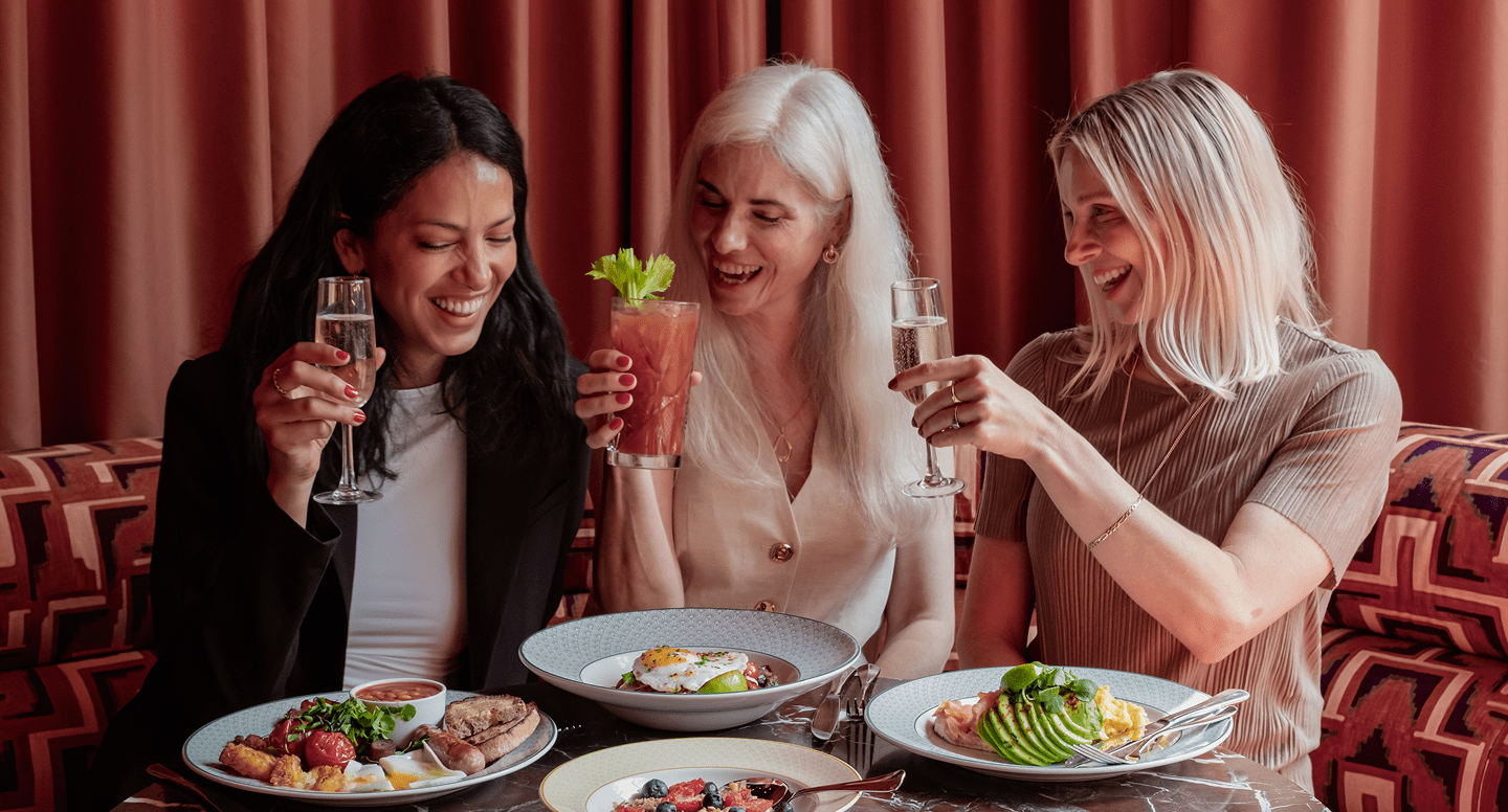Three women sitting together at a restaurant table, smiling and raising drinks in a toast. Plates of colorful, appetizing food are in front of them, and a curtain is in the background.
