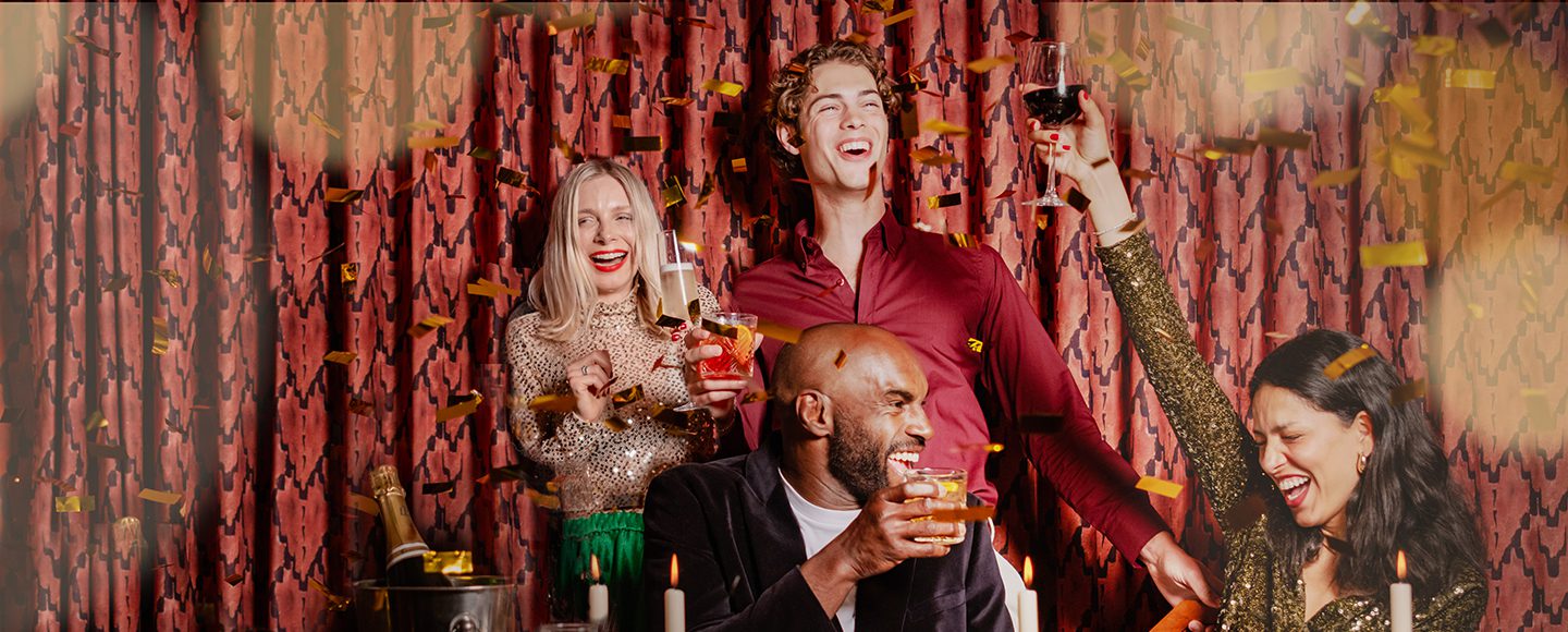 Four people dressed in festive outfits celebrate together, smiling and holding drinks, surrounded by falling gold confetti in front of a patterned curtain.