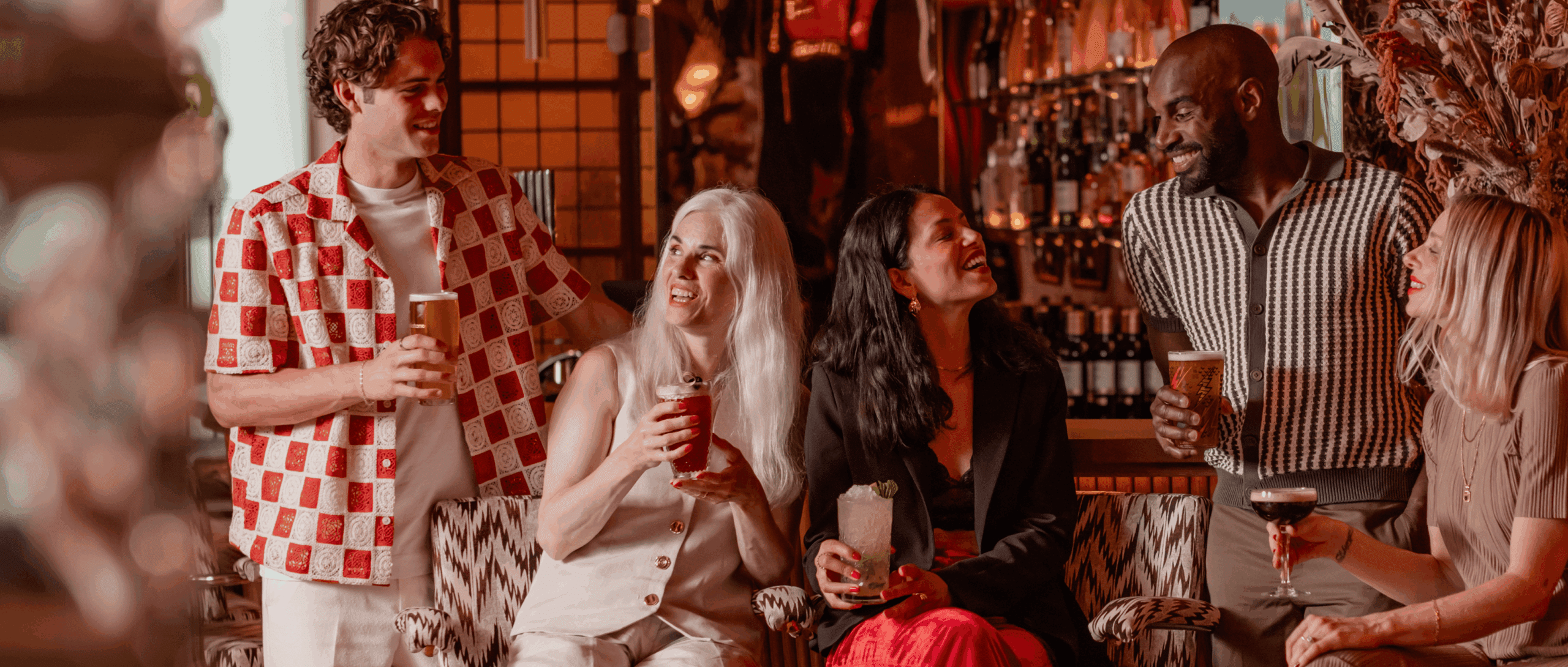 Five people sit together in a stylish bar, holding drinks and smiling as they enjoy each other's company. The setting is warmly lit, with bottles and decor visible in the background.