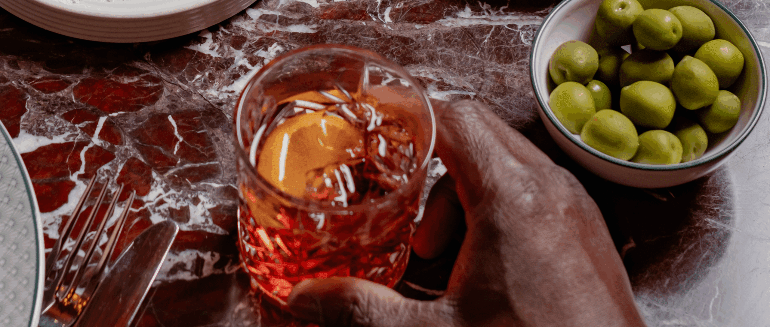 A hand holds a glass of amber-colored drink with ice on a marbled table, next to a bowl of green olives and a fork.