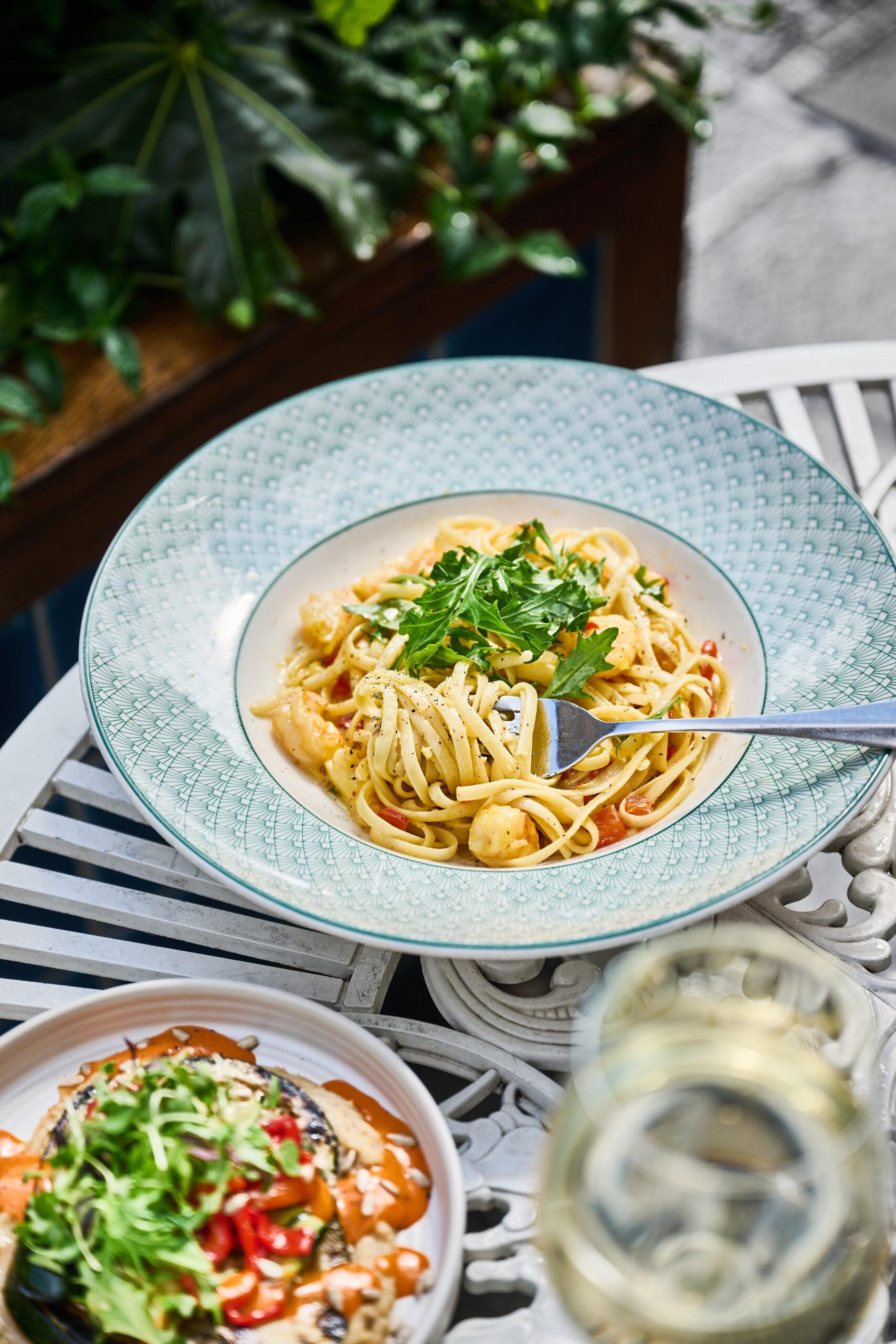 A plate of spaghetti garnished with fresh parsley sits on an outdoor table, with a fork twirled in the pasta. Nearby are a glass of white wine and a bowl of vegetables, with green plants in the background.
