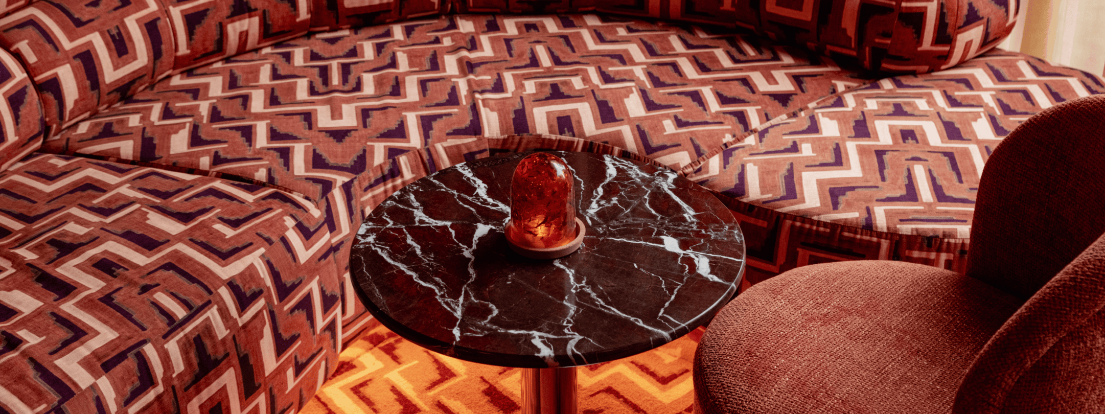 A round black marble coffee table with a small orange glass lamp sits between a curvy patterned sofa and a textured armchair in a retro-style living room.