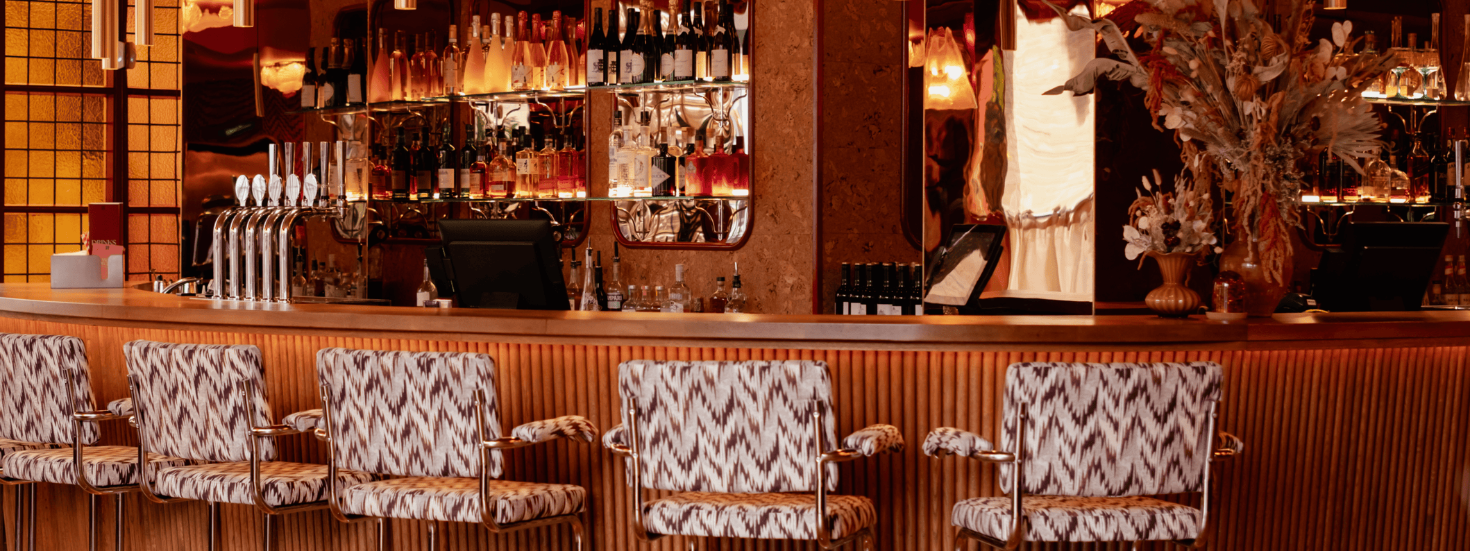 A stylish bar with patterned upholstered stools, a wooden counter, mirrored shelves displaying bottles and glasses, warm lighting, and a decorative vase with dried flowers.