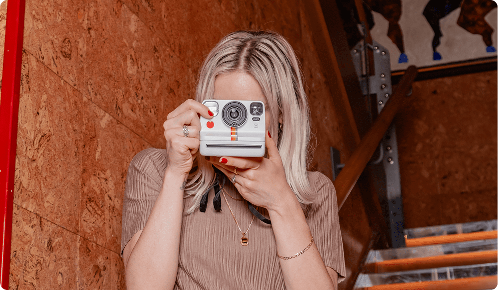 A person with blonde hair holds a vintage instant camera up to their face, preparing to take a photo while standing indoors near a wooden staircase.