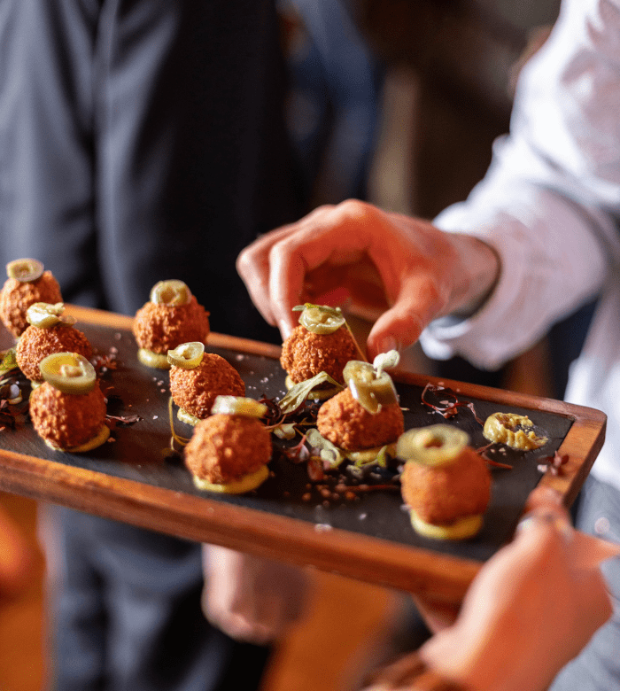 A person serves a wooden tray of bite-sized appetizers topped with sliced jalapeños and placed on small crackers, while others reach for the food. The background is blurred, focusing on the tray.