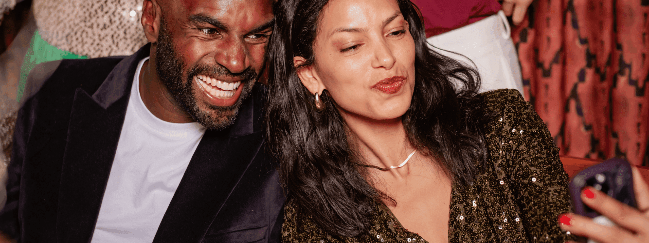 A smiling man and woman sit close together, taking a selfie. The woman wears a sparkly top and gold earrings, while the man wears a dark blazer. Both appear happy and relaxed in a festive setting.