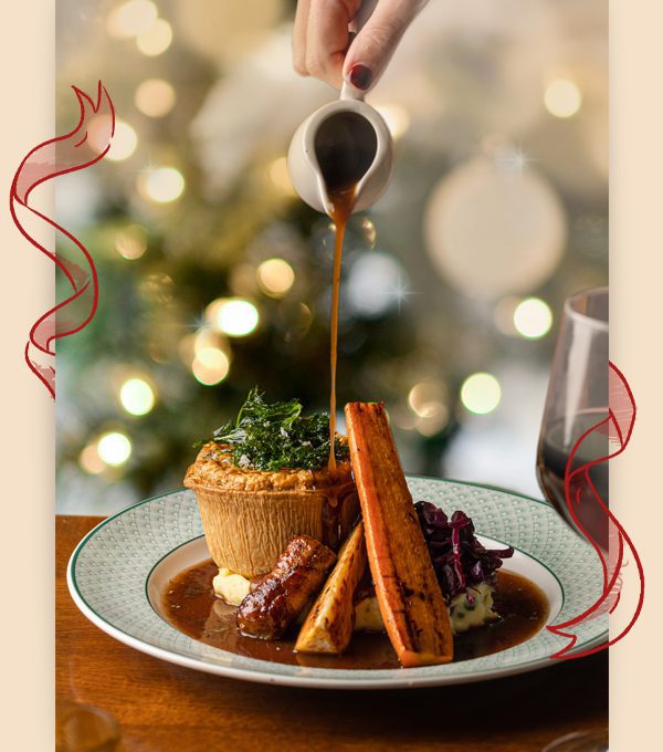 A hand pours gravy over a savory pie, roasted vegetables, and mashed potatoes on a plate. A festive, blurred background with lights and a glass of red wine enhance the cozy holiday atmosphere.