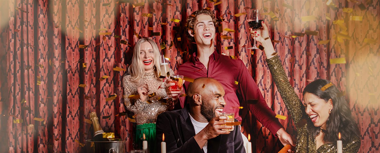 Four people dressed in festive attire celebrate with drinks, laughing and smiling as gold confetti falls around them. They are seated at a decorated table with candles against a patterned curtain backdrop.