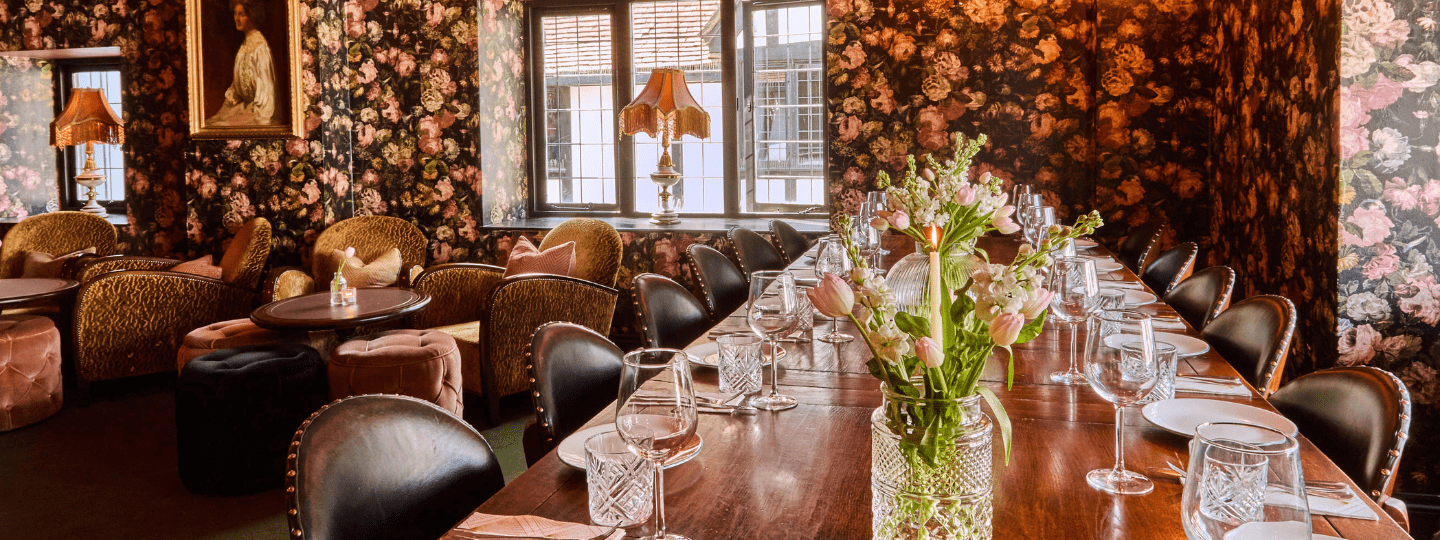 A long wooden dining table with elegant place settings and a vase of flowers is surrounded by velvet chairs in a room with floral wallpaper, vintage lamps, and large windows letting in natural light.