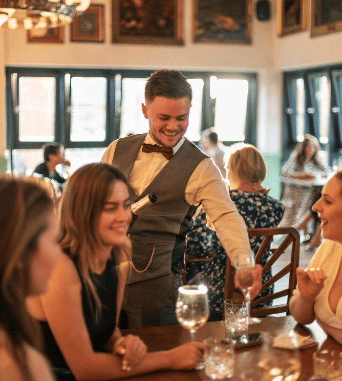 A smiling waiter in a vest and bow tie serves drinks to a group of women sitting at a wooden table in a bright, warmly lit restaurant. Other diners can be seen in the background.