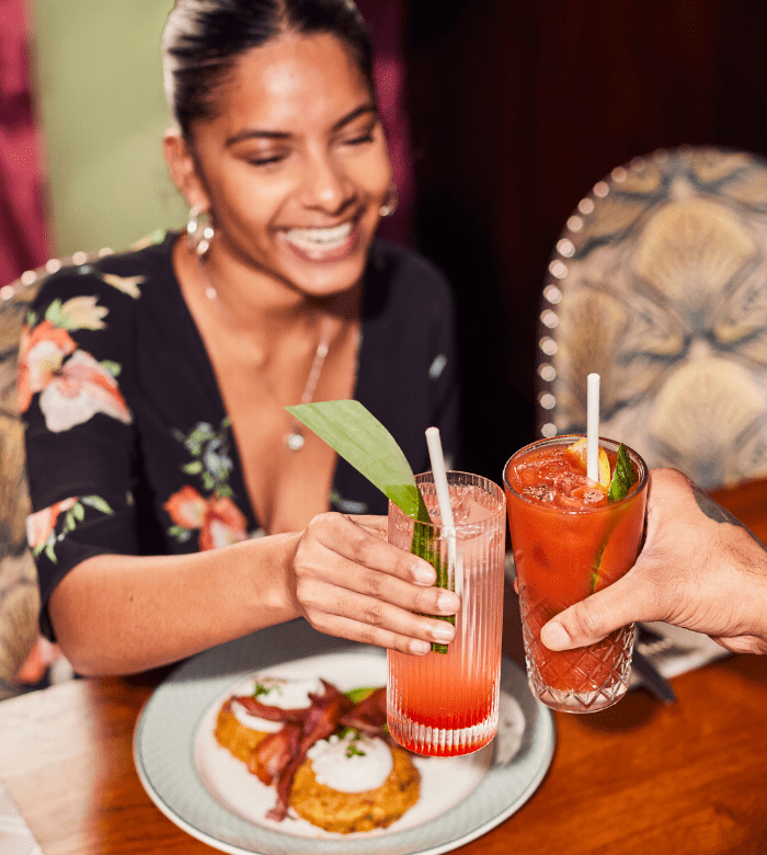 A smiling woman clinks glasses with another person at a restaurant table, holding colorful drinks. A plate of food with what appears to be bacon and an egg is in front of her. The setting is cozy and warmly lit.