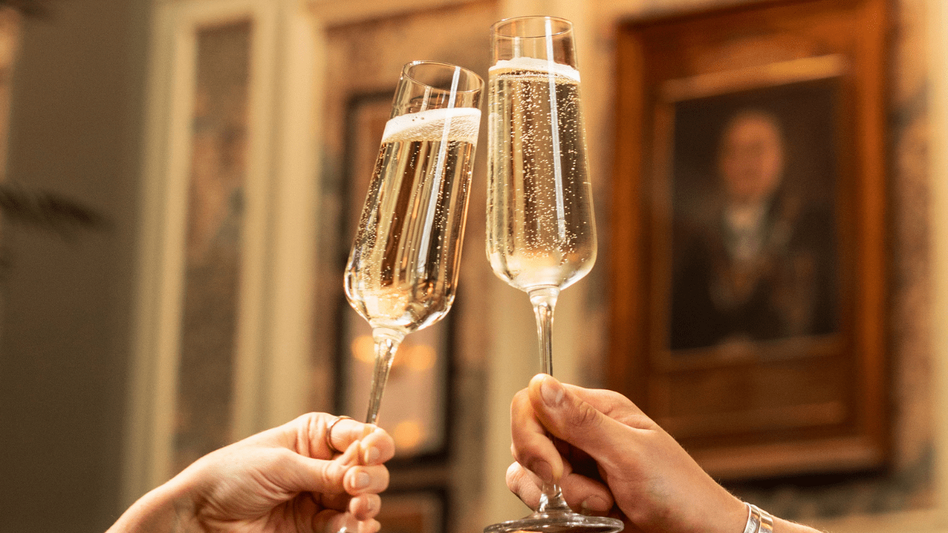 Two hands clinking champagne glasses filled with sparkling wine in a celebratory toast, with a blurred background featuring framed pictures on the wall.